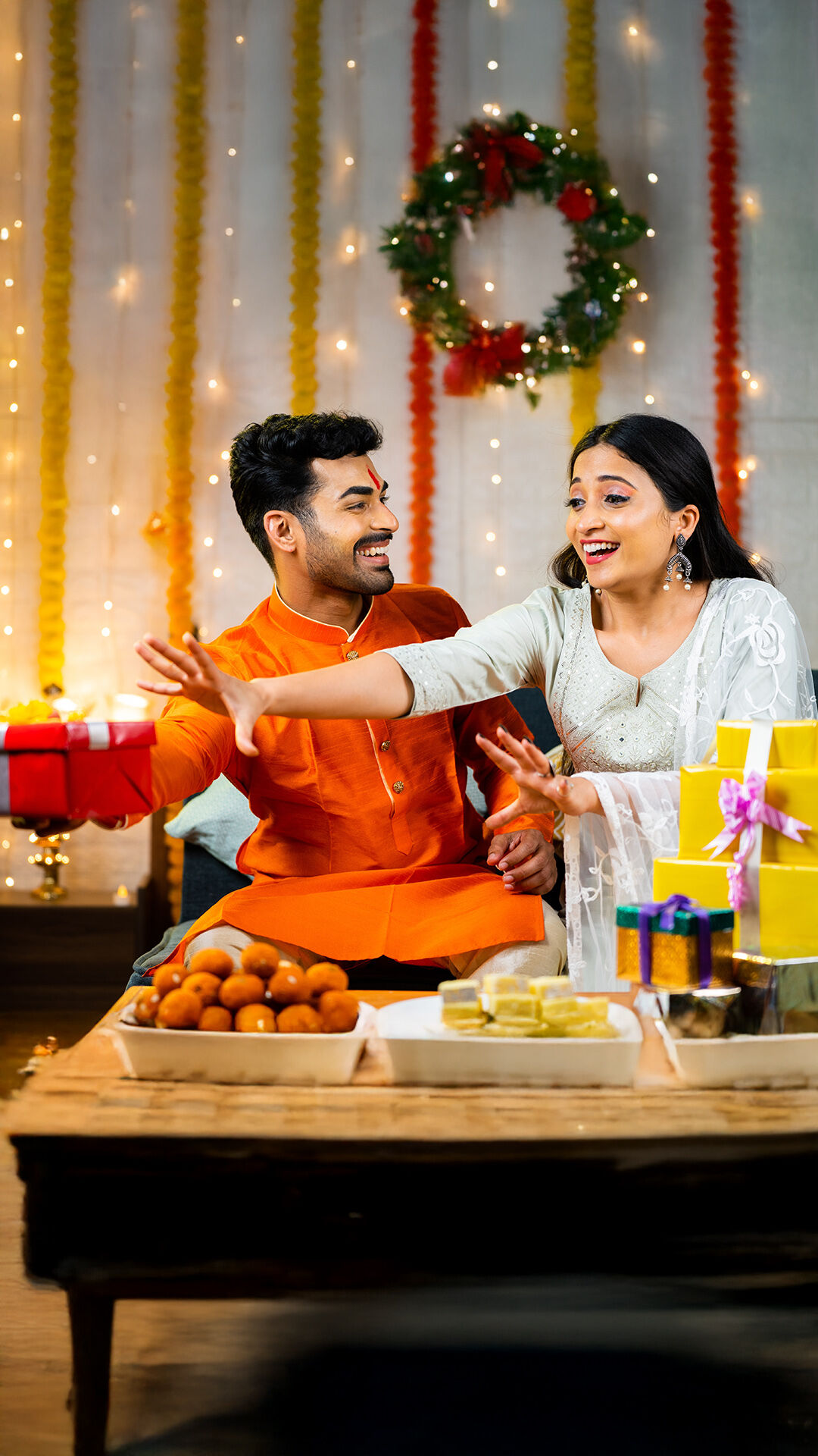 Indian couple playfully exchanging gifts during Diwali celebration, with traditional sweets, festive decor, and fairy lights in the background.